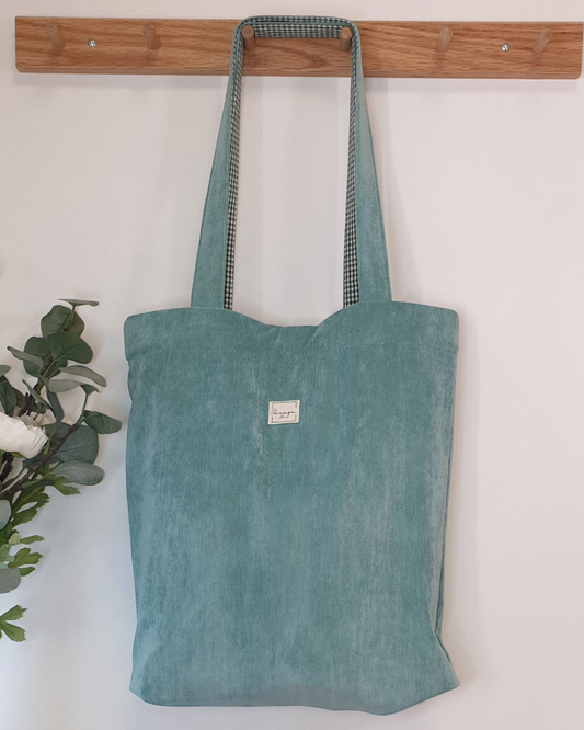 Tote bag artesanal SAVAGE LINE de pana fina color agua marina con forro interior de cuadros vichy colgado en pared